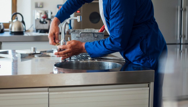 A technician from TCI Plumbing, which offers residential plumbing services, works on a kitchen sink with his tool kit in the background.